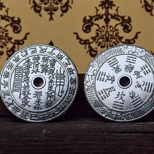 May include: Two silver-colored, round coins with central holes and intricate Chinese characters. The coins are displayed on a dark wooden surface against a patterned background. The coins' detailed designs suggest they are collectible or historical items.