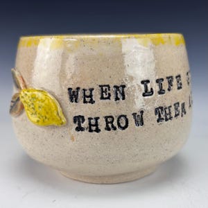 May include: A ceramic mug with a speckled beige finish and a yellow rim. The mug features a yellow lemon design and the text "WHEN LIFE THROWS THEM".