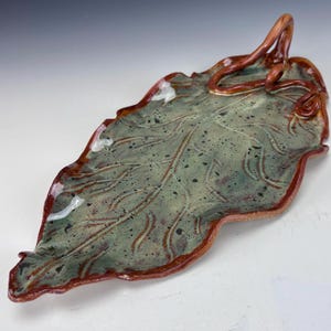 May include: A ceramic leaf-shaped serving dish. The dish is primarily green with brown accents and a textured surface. The rim and handle are brown. The leaf design features detailed veining and a curled handle.