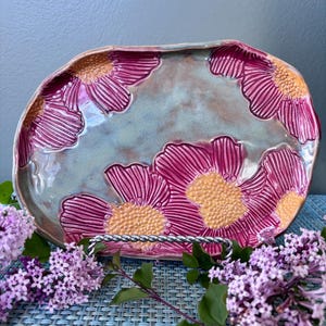 May include: A ceramic serving platter with a light blue-green base and raised pink flower designs with yellow centers. The platter is displayed on a silver metal stand, with purple flowers in the foreground.