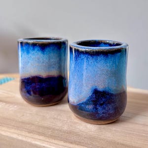 May include: Two handmade ceramic tumblers with a gradient of blue hues, transitioning from a dark indigo base to a lighter sky blue at the top. The rims have a dark brown glaze. The tumblers are on a wooden surface.