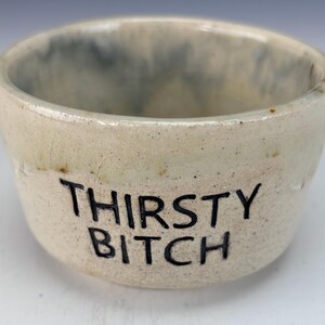 May include: A small, off-white ceramic bowl with the words "THIRSTY BITCH" in black lettering. The bowl has a slightly speckled texture and a blue-grey interior. The bowl is round and has a simple, rustic design.
