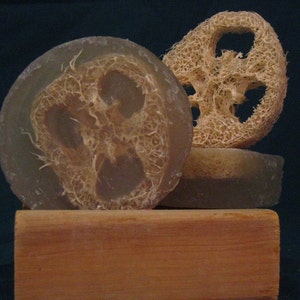May include: A close-up of natural soap bars with a loofah sponge. The soap is round and translucent, with a visible loofah center. The loofah sponge is a light beige color, and the soap is resting on a wooden block.