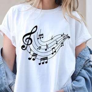 May include: White t-shirt featuring a black treble clef and musical notes design. The musical notes flow across the shirt in a curved pattern. The shirt is made of a soft, comfortable material.