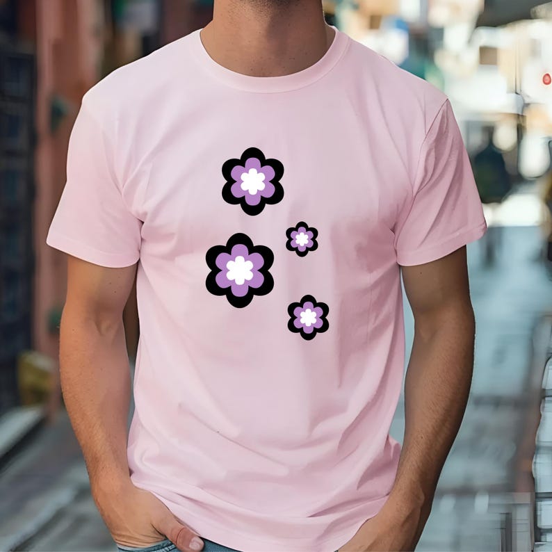 May include: A pale pink t-shirt with a floral pattern. The flowers have black outlines, purple petals, and white centres. The design is repeated in different sizes across the shirt. The t-shirt is short-sleeved.