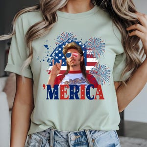 Op de afbeelding: Lichtgroen t-shirt met een afbeelding van een man met een zonnebril en een hanenkam, met het woord 'MERICA in rood, wit en blauw. Het ontwerp bevat een Amerikaanse vlag en vuurwerk.
