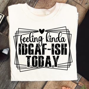 K&ouml;nnte beinhalten: Wei&szlig;es T-Shirt mit dem schwarzen Schriftzug &bdquo;feeling kinda IDGAF-ISH TODAY&ldquo; in einem geometrischen Rahmen. Ein kleines schwarzes Herz befindet sich &uuml;ber dem Wort &bdquo;feeling&ldquo;. Das T-Shirt liegt auf einer Holzoberfl&auml;che.