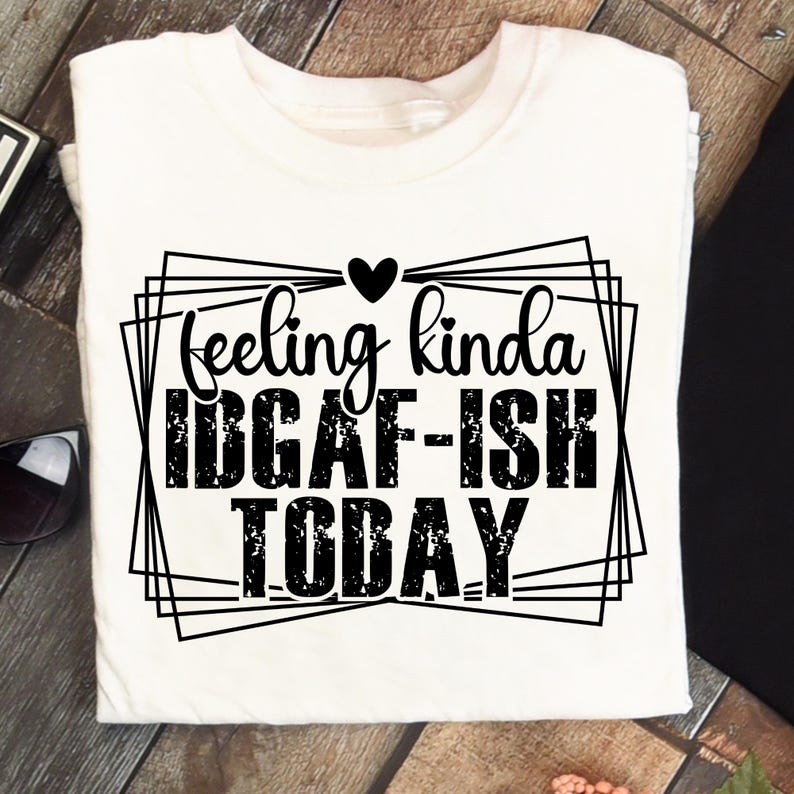 K&ouml;nnte beinhalten: Wei&szlig;es T-Shirt mit dem schwarzen Schriftzug &bdquo;feeling kinda IDGAF-ISH TODAY&ldquo; in einem geometrischen Rahmen. Ein kleines schwarzes Herz befindet sich &uuml;ber dem Wort &bdquo;feeling&ldquo;. Das T-Shirt liegt auf einer Holzoberfl&auml;che.