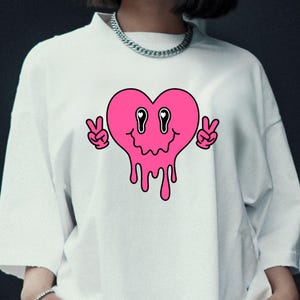 May include: White t-shirt with a pink, melting heart design and a smiling face. The heart has two peace signs on either side. The shirt has a crew neck and short sleeves.