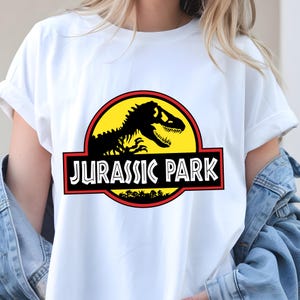 May include: White t-shirt featuring the Jurassic Park logo. The logo includes a black dinosaur skeleton silhouette on a yellow circle, with a red border and the words "JURASSIC PARK" in white.