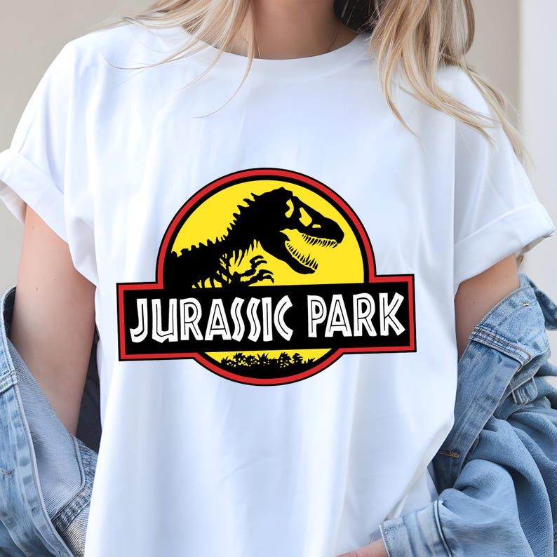 May include: White t-shirt featuring the Jurassic Park logo. The logo includes a black dinosaur skeleton silhouette on a yellow circle, with a red border and the words "JURASSIC PARK" in white.