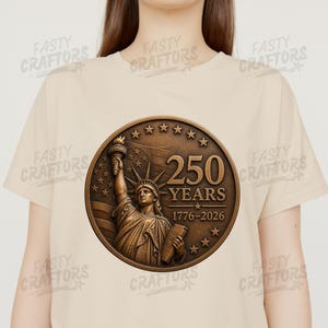 K&ouml;nnte beinhalten: Ein beiges T-Shirt mit einer kreisf&ouml;rmigen, bronzefarbenen Grafik der Freiheitsstatue. Das Design enth&auml;lt den Text 250 YEARS und 1776-2026, umgeben von Sternen und einem schwachen amerikanischen Flaggenhintergrund zur Feier des 250-j&auml;hrigen Jubil&auml;ums.