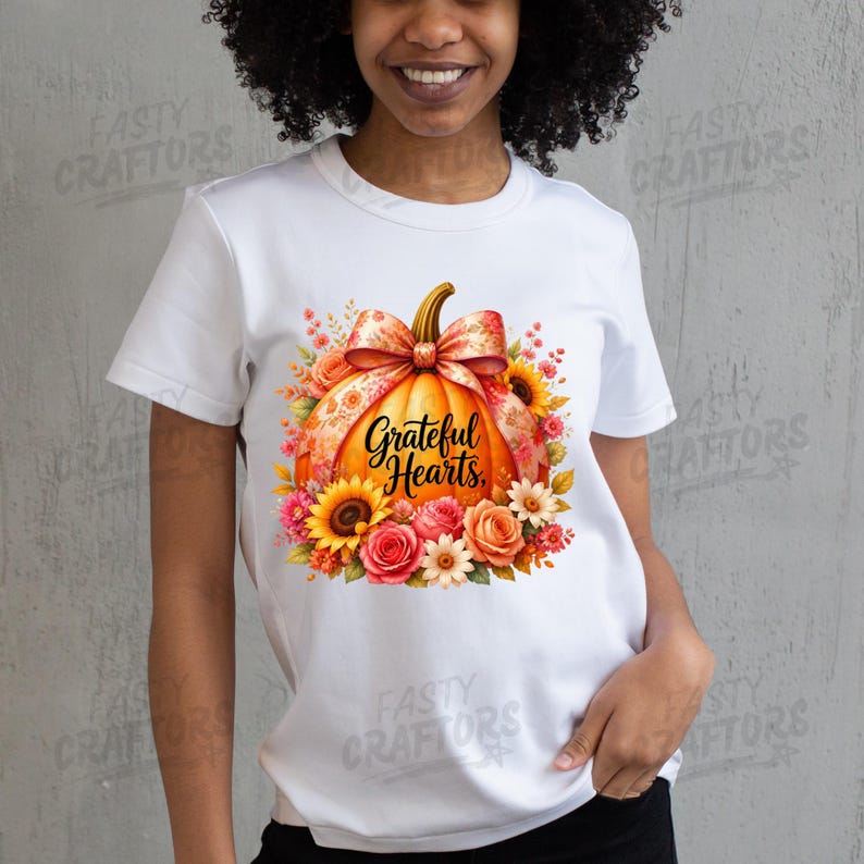 May include: A white short-sleeved t-shirt featuring a central graphic of an orange pumpkin adorned with a patterned bow and surrounded by colourful sunflowers and roses. The text Grateful Hearts is printed in black script across the pumpkin.