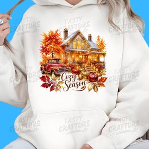 May include: A cream-coloured hooded sweatshirt featuring a graphic print of a house with autumn foliage, a red pickup truck, and pumpkins. The text "Cozy Season" is printed in a brown script font below the house, surrounded by colourful fall leaves.
