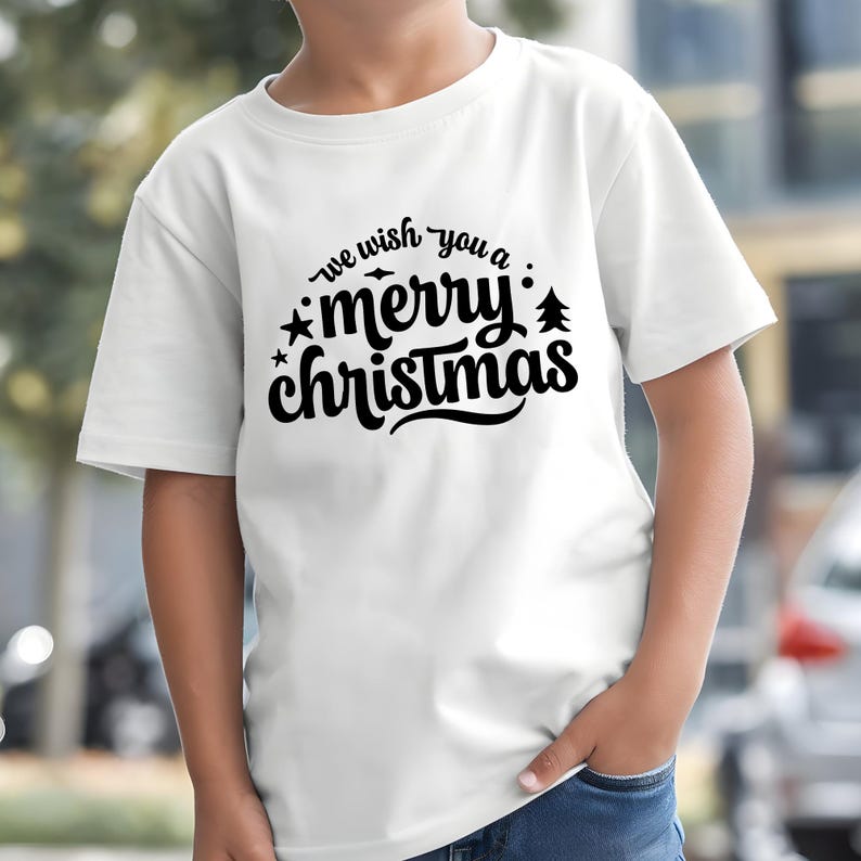 May include: White t-shirt with the text "we wish you a merry Christmas" in black script. The design includes stars and a small Christmas tree. The shirt is short-sleeved and appears to be made of cotton.