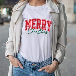 Puede incluir: Camiseta blanca con la frase "MERRY Christmas" en letras rojas y verdes. La palabra "MERRY" est&aacute; en negrita, con un estilo universitario, mientras que "Christmas" est&aacute; en cursiva. La camiseta se lleva con vaqueros azules y una chaqueta de color claro.