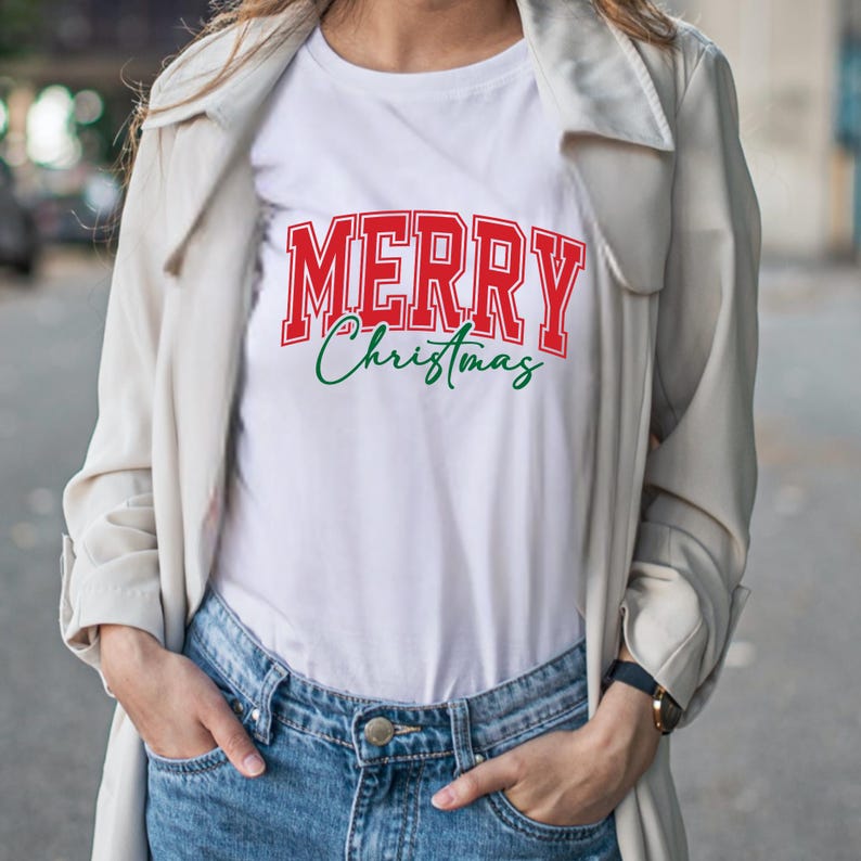 Puede incluir: Camiseta blanca con la frase "MERRY Christmas" en letras rojas y verdes. La palabra "MERRY" est&aacute; en negrita, con un estilo universitario, mientras que "Christmas" est&aacute; en cursiva. La camiseta se lleva con vaqueros azules y una chaqueta de color claro.