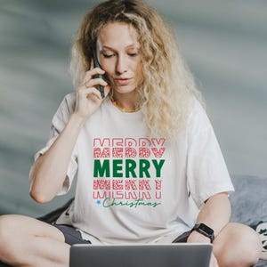 Puede incluir: Camiseta blanca con un dise&ntilde;o festivo "MERRY CHRISTMAS" en rojo y verde. El texto est&aacute; dispuesto en formato apilado con patrones decorativos. La camiseta la lleva una persona.