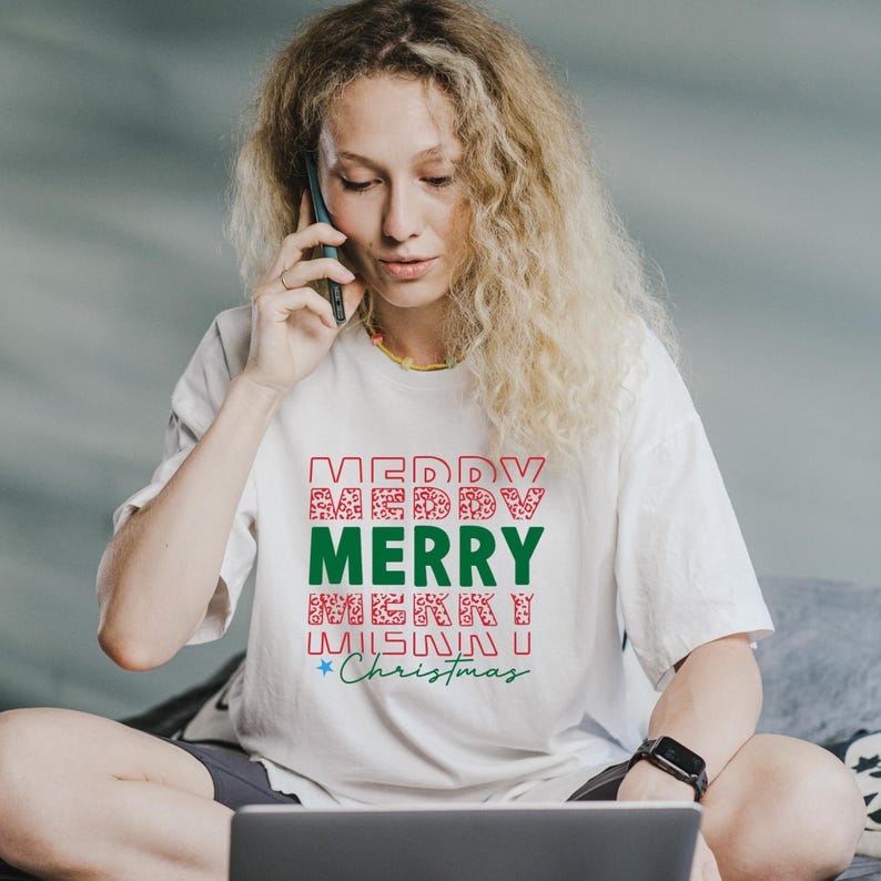 Puede incluir: Camiseta blanca con un dise&ntilde;o festivo "MERRY CHRISTMAS" en rojo y verde. El texto est&aacute; dispuesto en formato apilado con patrones decorativos. La camiseta la lleva una persona.