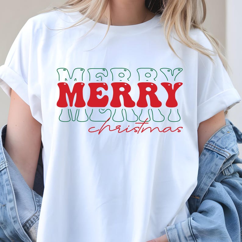Puede incluir: Camiseta blanca con las palabras "MERRY" en rojo, superpuestas a "christmas" en escritura roja. La palabra "MERRY" tambi&eacute;n est&aacute; escrita en verde encima y debajo del texto rojo. La camiseta tiene mangas cortas y un corte holgado.
