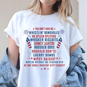 Op de afbeelding: Wit T-shirt met rode, witte en blauwe tekst en afbeeldingen. De tekst bevat zinnen als "Whistlin' Bungholes" en "Cherry Bombs". Het ontwerp bevat vuurwerk en steraccenten, wat een patriottisch thema cre&euml;ert.