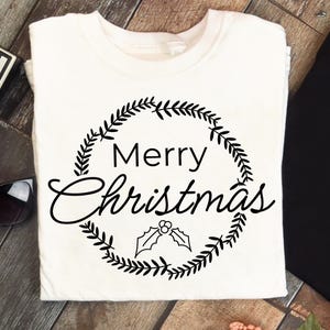 May include: White t-shirt with a black wreath design and the words "Merry Christmas" in a script font. The wreath includes leaves and holly berries, perfect for the holiday season. A festive apparel item.