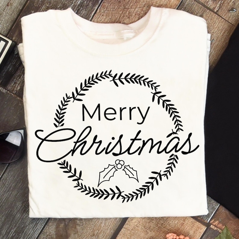 May include: White t-shirt with a black wreath design and the words "Merry Christmas" in a script font. The wreath includes leaves and holly berries, perfect for the holiday season. A festive apparel item.