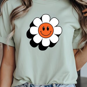May include: A pale green t-shirt with a large daisy graphic. The daisy has white petals, an orange centre with a smiling face, and a black outline. The shirt is made of a soft material.