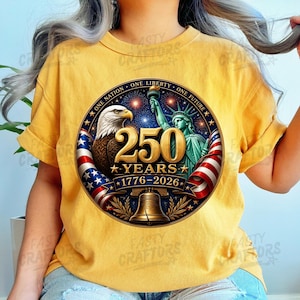 K&ouml;nnte beinhalten: Ein senfgelbes T-Shirt mit einer kreisf&ouml;rmigen Grafik, die einen Wei&szlig;kopfseeadler, die Freiheitsstatue, eine Glocke und US-Flaggen zeigt. Der Text lautet 250 YEARS 1776-2026 und ONE NATION ONE LIBERTY ONE FUTURE mit Feuerwerk im Hintergrund.
