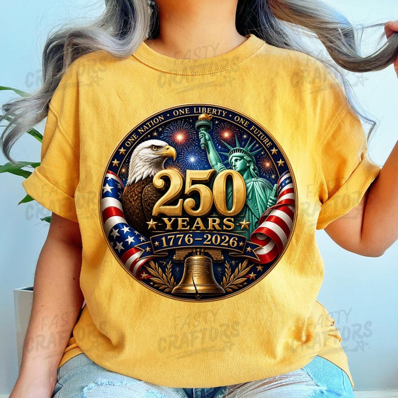 K&ouml;nnte beinhalten: Ein senfgelbes T-Shirt mit einer kreisf&ouml;rmigen Grafik, die einen Wei&szlig;kopfseeadler, die Freiheitsstatue, eine Glocke und US-Flaggen zeigt. Der Text lautet 250 YEARS 1776-2026 und ONE NATION ONE LIBERTY ONE FUTURE mit Feuerwerk im Hintergrund.