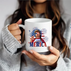 Op de afbeelding: Witte keramische mok met een patriottisch ontwerp. De mok toont een afbeelding van een man met een zonnebril en het woord 'Merica in rood en blauw. Het ontwerp bevat een Amerikaanse vlag en vuurwerk.