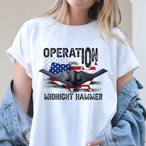 Pode incluir: Camiseta branca com um design de bombardeiro furtivo preto sobre um mapa dos Estados Unidos e a bandeira americana. O texto "OPERATION" e "MIDNIGHT HAMMER" são impressos acima e abaixo do gráfico.