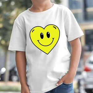 May include: White t-shirt with a large yellow heart design. The heart has a smiling face with black eyes and a black outline. The shirt is short-sleeved and made of a light fabric.