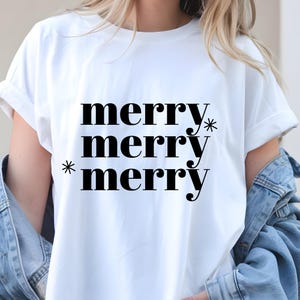 May include: White t-shirt with the word "merry" repeated three times in a bold, black font. Asterisks are placed next to the first and third instances of the word. The shirt is a casual style with rolled-up sleeves.