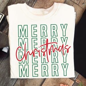 Puede incluir: Camiseta blanca con las palabras "MERRY Christmas" en letras verdes y rojas. La palabra "MERRY" est&aacute; apilada verticalmente cuatro veces, con "Christmas" escrito en cursiva sobre las instancias tercera y cuarta de "MERRY".