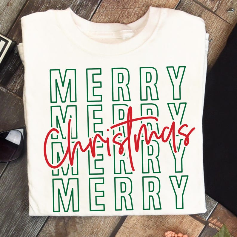Puede incluir: Camiseta blanca con las palabras "MERRY Christmas" en letras verdes y rojas. La palabra "MERRY" est&aacute; apilada verticalmente cuatro veces, con "Christmas" escrito en cursiva sobre las instancias tercera y cuarta de "MERRY".