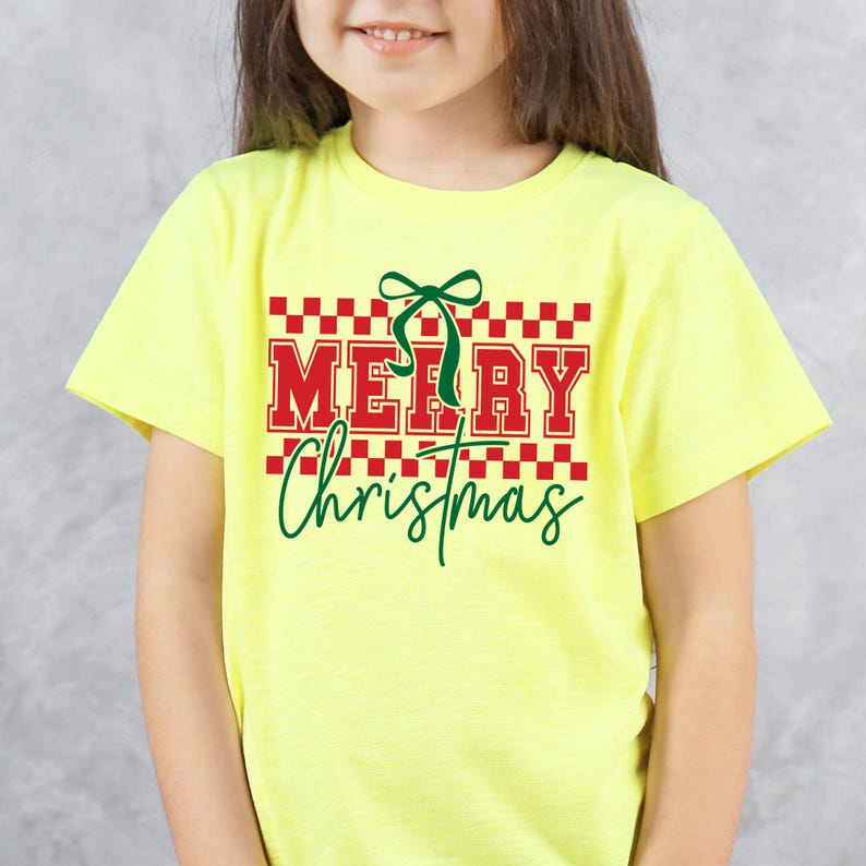Puede incluir: Una camiseta amarilla de manga corta con las palabras "MERRY Christmas" en rojo y verde. El texto est&aacute; sobre un patr&oacute;n de cuadros rojos y blancos, con un lazo verde encima de la palabra "MERRY".