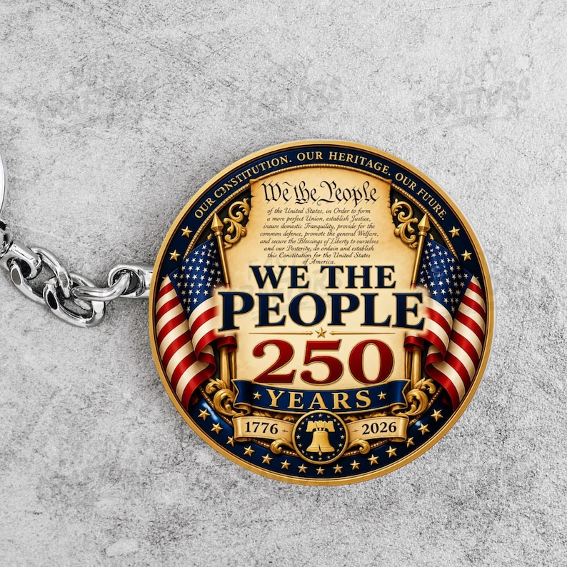 K&ouml;nnte beinhalten: Ein runder Schl&uuml;sselanh&auml;nger mit goldenem Rand und zwei amerikanischen Flaggen, die eine zentrale Grafik im Pergamentstil flankieren. Der Text lautet We The People, 250 Years, 1776-2026, und Our Constitution, Our Heritage, Our Future.