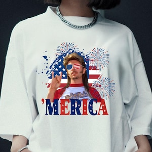 Op de afbeelding: Wit T-shirt met een afbeelding van een man met een zonnebril in de stijl van de Amerikaanse vlag, een hanenkam en het woord "'MERICA" in rood, wit en blauw. Het ontwerp bevat vuurwerk en een Amerikaanse vlag achtergrond.