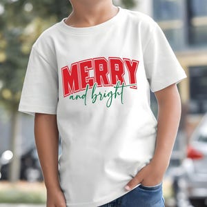Puede incluir: Camiseta blanca con las palabras "MERRY and bright" en letras rojas y verdes. La palabra "MERRY" est&aacute; en una fuente en negrita y en bloque, mientras que "and bright" est&aacute; en cursiva.