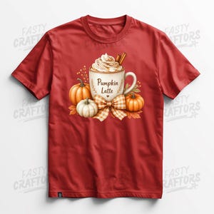 May include: A red short-sleeved t-shirt featuring a graphic of a mug filled with a pumpkin latte, topped with whipped cream and cinnamon sticks. The mug is labelled Pumpkin Latte and is surrounded by orange pumpkins and a plaid bow.