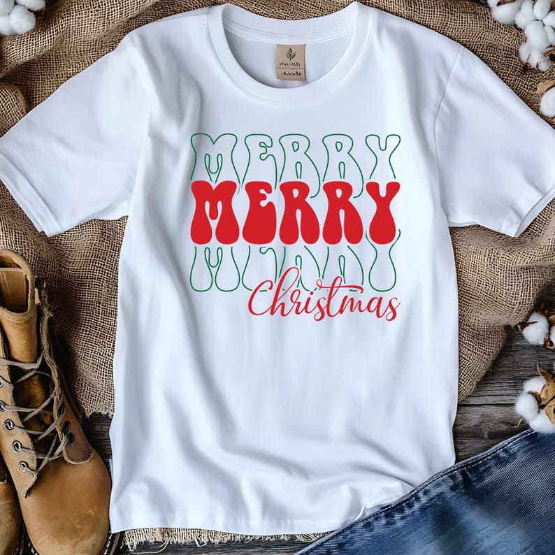 Puede incluir: Camiseta blanca con las palabras "MERRY MERRY Christmas" en una fuente retro. La palabra "MERRY" es roja, con contorno verde. "Christmas" es rojo. La camiseta est&aacute; hecha de un material suave.