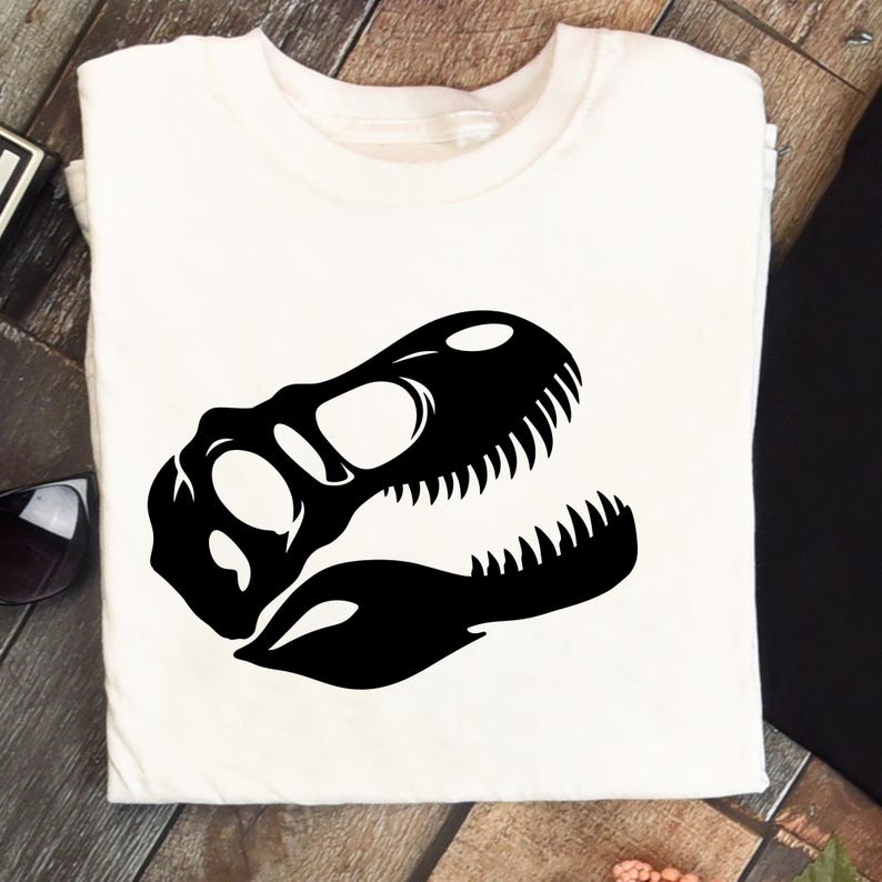 May include: White t-shirt featuring a black dinosaur skull graphic. The design shows the skull's outline, including the eye sockets and teeth. The shirt is folded, with the graphic centered on the front.