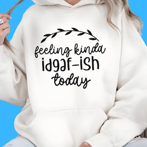 K&ouml;nnte beinhalten: Cremefarbener Kapuzenpullover mit schwarzem Aufdruck "feeling kinda id9af-ish today" und einem schwarzen Blattdesign. Der Hoodie hat eine Vordertasche und eine Kapuze.