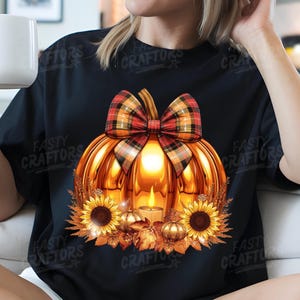 May include: A black t-shirt featuring a graphic of a large, glowing orange pumpkin topped with a red and black plaid bow. The pumpkin contains a lit candle and is surrounded by sunflowers, smaller golden pumpkins, and autumn leaves.