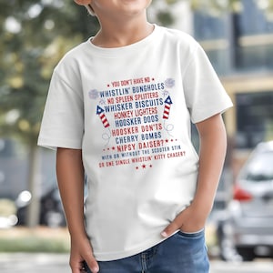Op de afbeelding: Een wit T-shirt met een patriottisch ontwerp met rode en blauwe tekst. De tekst bevat zinnen als "Whistlin' Bungholes" en "Cherry Bombs". Het T-shirt wordt door een kind gedragen.