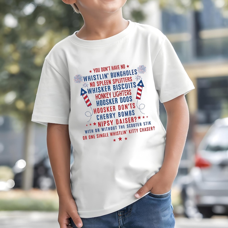 Op de afbeelding: Een wit T-shirt met een patriottisch ontwerp met rode en blauwe tekst. De tekst bevat zinnen als "Whistlin' Bungholes" en "Cherry Bombs". Het T-shirt wordt door een kind gedragen.