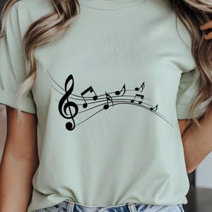 May include: A light green t-shirt featuring a black musical staff with a treble clef and musical notes. The design is centered on the front of the shirt. The shirt is made of a soft material.
