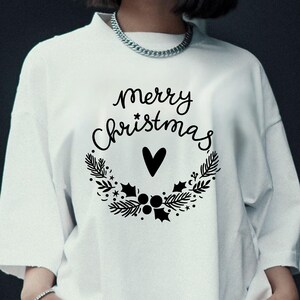 May include: White t-shirt with a black wreath design and the words "Merry Christmas" in a script font. A black heart is in the center of the wreath. The shirt is a casual style.