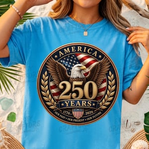 K&ouml;nnte beinhalten: Ein hellblaues T-Shirt mit einer kreisf&ouml;rmigen Grafik, die einen Wei&szlig;kopfseeadler, die amerikanische Flagge und den Text AMERICA 250 YEARS 1776-2026 OUR PAST OUR FUTURE zeigt. Das Design enth&auml;lt goldene Lorbeerzweige und ein gestreiftes Wappen.